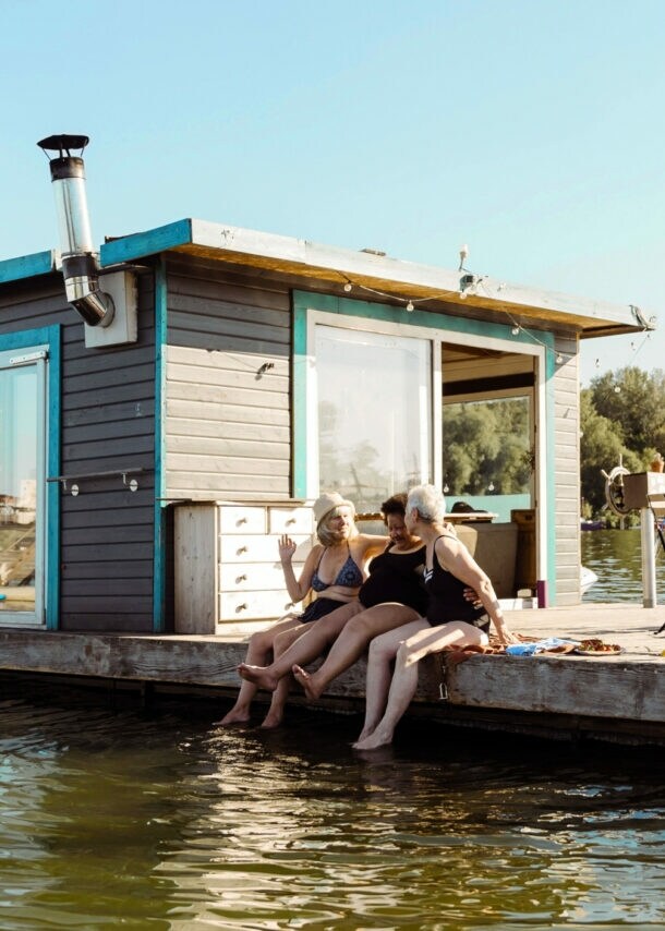 Drei Frauen in Badekleidung sitzen am Rand eines Pontons mit Hausboot und lassen ihre Füße ins Wasser baumeln bei Sonnenschein.