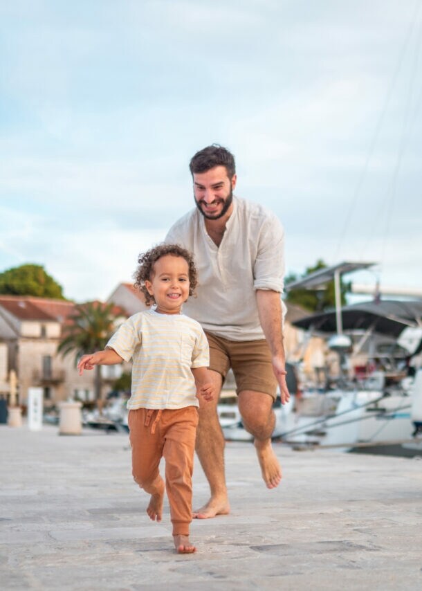 Ein lächelnder Mann läuft barfuß einem fröhlichen kleinen Kind auf einem gepflasterten Hafenweg mit Segelbooten im Hintergrund nach