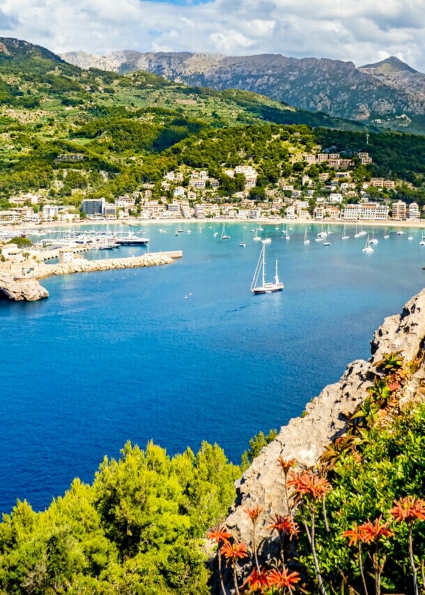 Bucht mit klarem blauem Wasser, Segelboot und Yachten, umgeben von grünen Hügeln und Bergen unter bewölktem Himmel