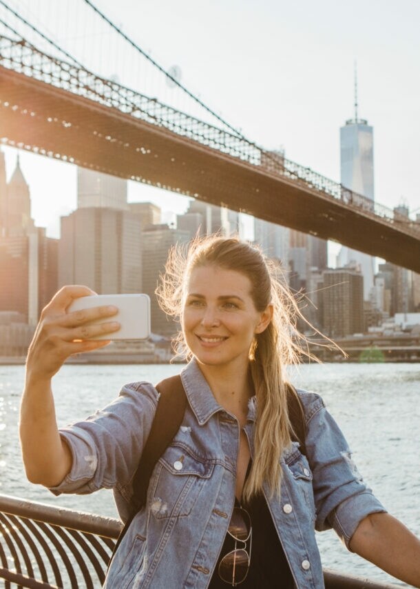 Frau macht Selfie vor der Brooklyn Bridge und der Skyline von New York bei Sonnenuntergang