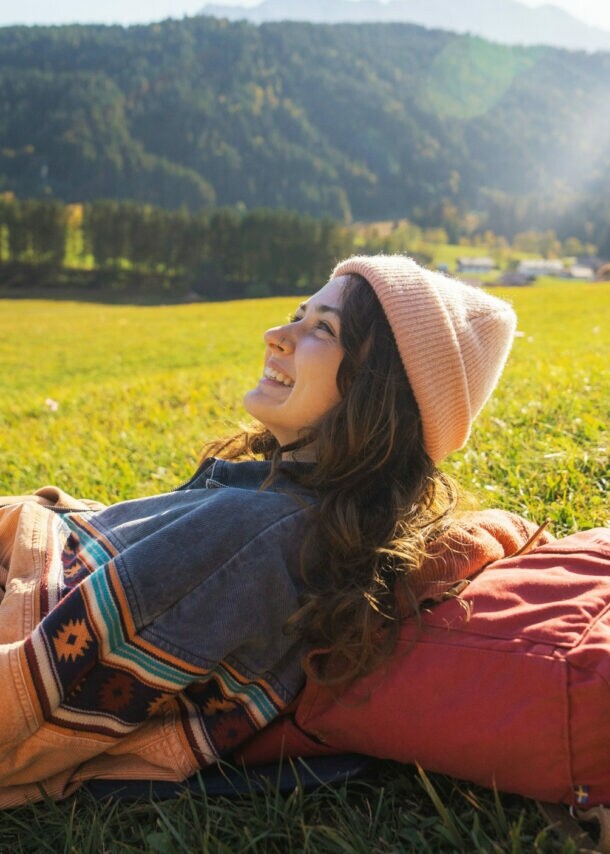 Eine Frau ruht sich auf einer Wiese in der Sonne aus, dabei liegt sie auf ihrem Wanderrucksack.