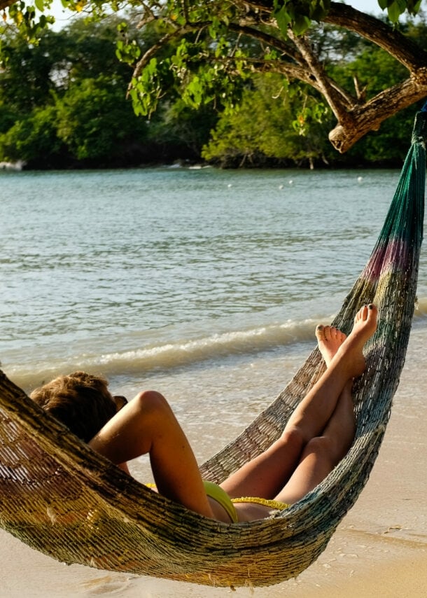 Person entspannt in einer Hängematte am Strand unter einem Baum mit Blick auf das Meer