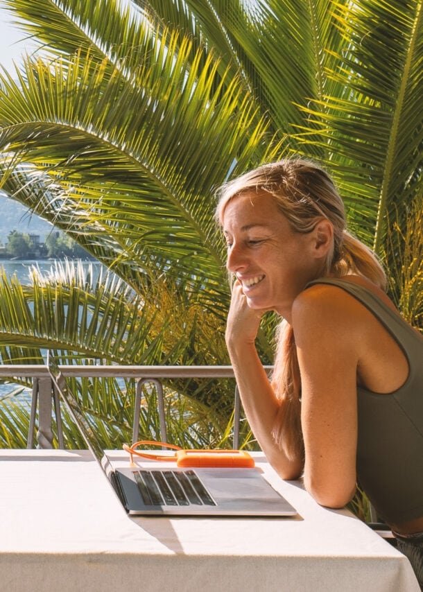 Eine Frau arbeitet am Laptop auf der Terrasse unter Palmen.