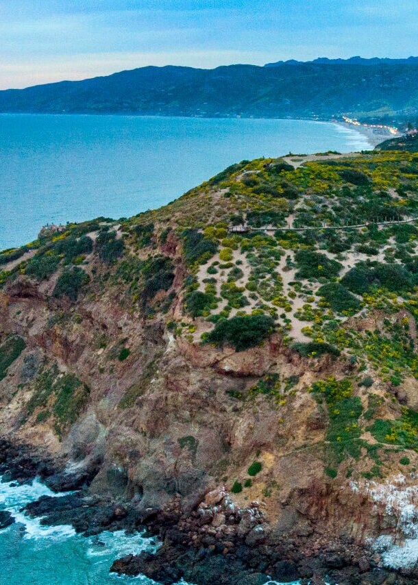 Klippen mit grüner Vegetation an der Küste bei Sonnenuntergang, dahinter Berge und ein Dorf.