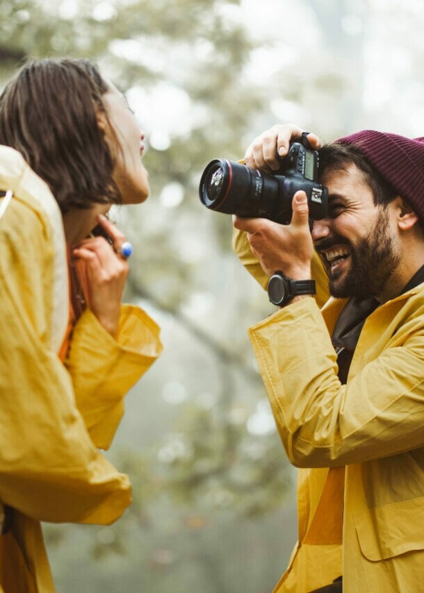 Mann mit roter Mütze und gelbem Regenmantel fotografiert lachende Frau mit blauem Rucksack und gelbem Regenmantel im Wald