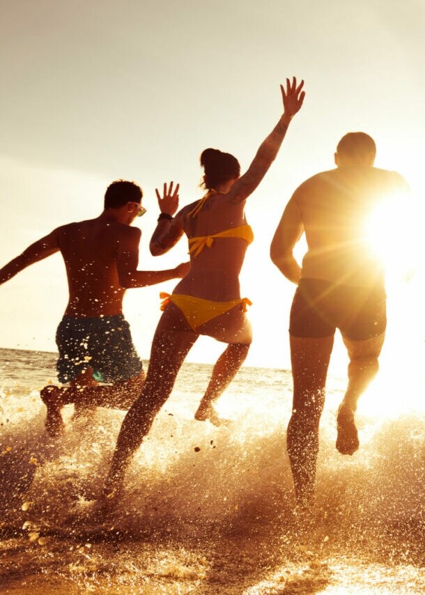 Gruppe von sechs jungen Menschen läuft im Sonnenuntergang ins Meer, Wasser spritzt um sie herum.