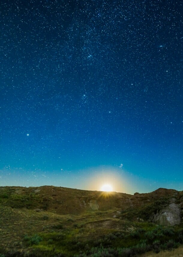 Klarer, tiefblauer Sternenhimmel mit aufgehendem Mond über einer felsigen Graslandschaft.
