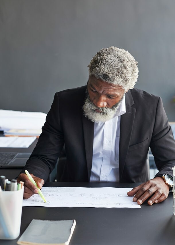 Ein älterer Mann arbeitet fokussiert im Büro an einer technischen Zeichnung.
