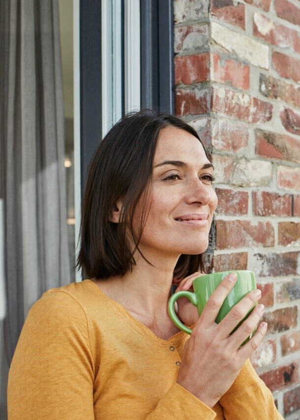 Eine Frau mit einer Tasse in der Hand lehnt entspannt an der Hauswand.
