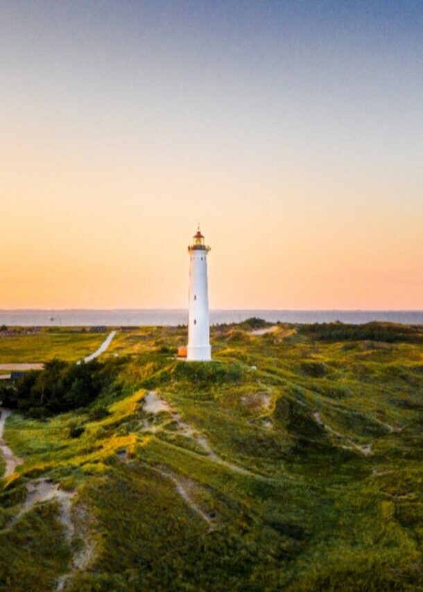 Weißer Leuchtturm auf grünen Dünen bei Sonnenuntergang mit Dorf und Meer im Hintergrund.