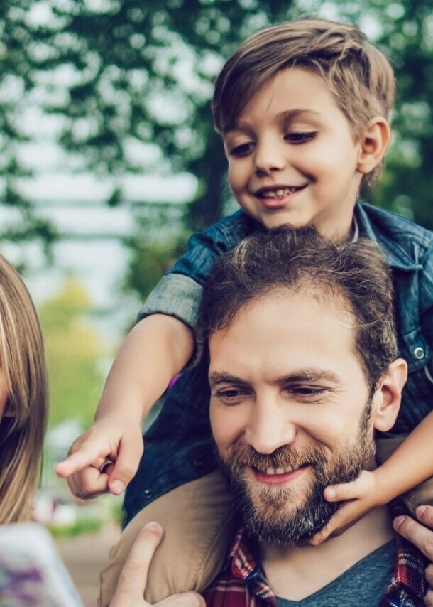 Vater trägt Sohn auf Schultern, Mutter hält Karte, sie stehen in einem Park mit Bäumen und Gebäuden im Hintergrund
