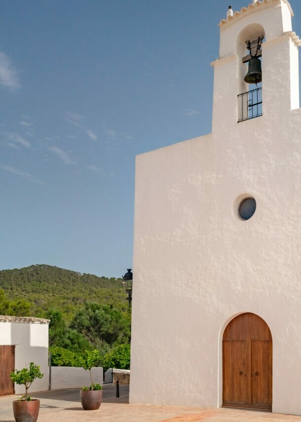Weiße Kirche mit Glockenturm und drei Kreuzen an der Wand, umgeben von Gebäuden und Bergen unter blauem Himmel
