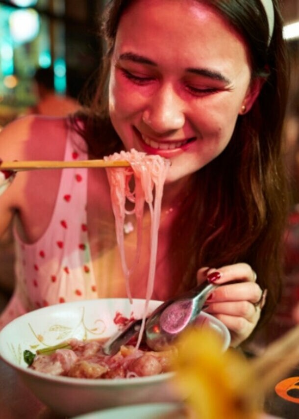 Nahaufnahme einer jungen, lächelnden Frau beim Essen eines asiatischen Nudelgerichtes mit Essstäbchen auf einem Straßenmarkt bei Nacht.