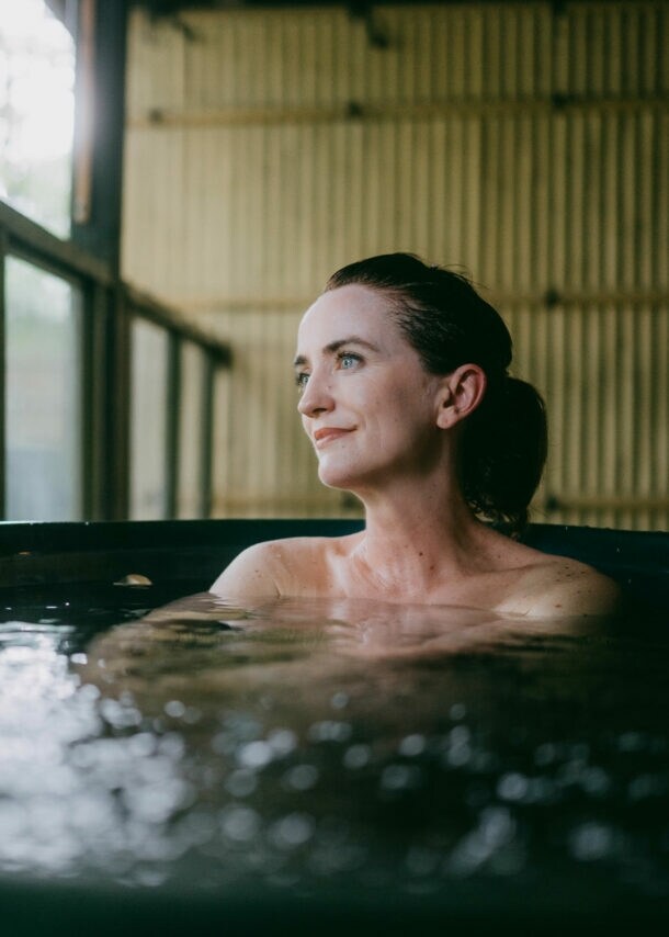 Eine Frau sitzt in einem Thermalbad aus dunklem Stein in einem japanischen Spa mit Bambuswand und schaut zufrieden durch ein Fenster hinaus.