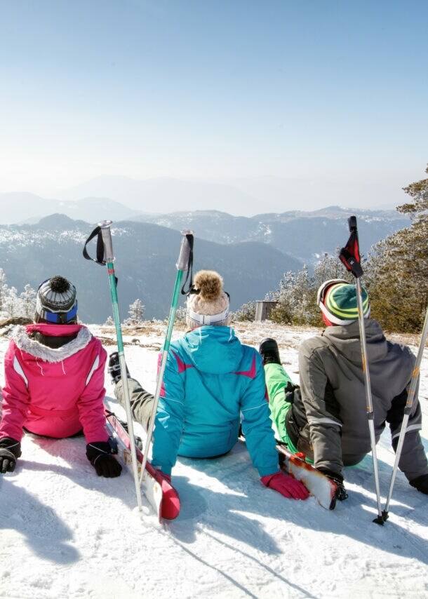 Fünf Personen mit Snowboards und Ski sitzen mit Blick auf eine weitere Berglandschaft auf einem Berg im Schnee.