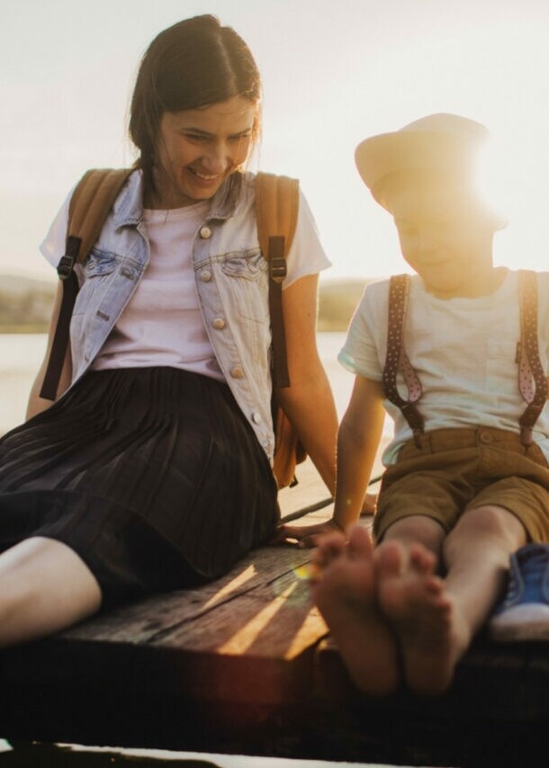 Mutter und Kind sitzen barfuß auf einem Holzsteg am See, die Sonne scheint im Hintergrund