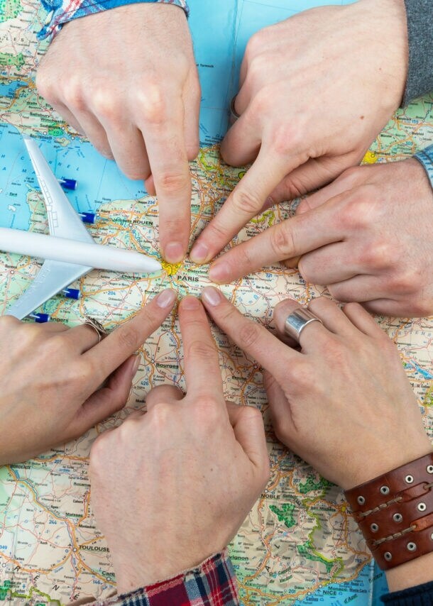 Sechs Personen, von denen nur die Hände zu sehen sind, zeigen auf einen Punkt auf einer Landkarte. Daneben liegen ein alter Fotoapparat und ein Modell eines Flugzeugs.