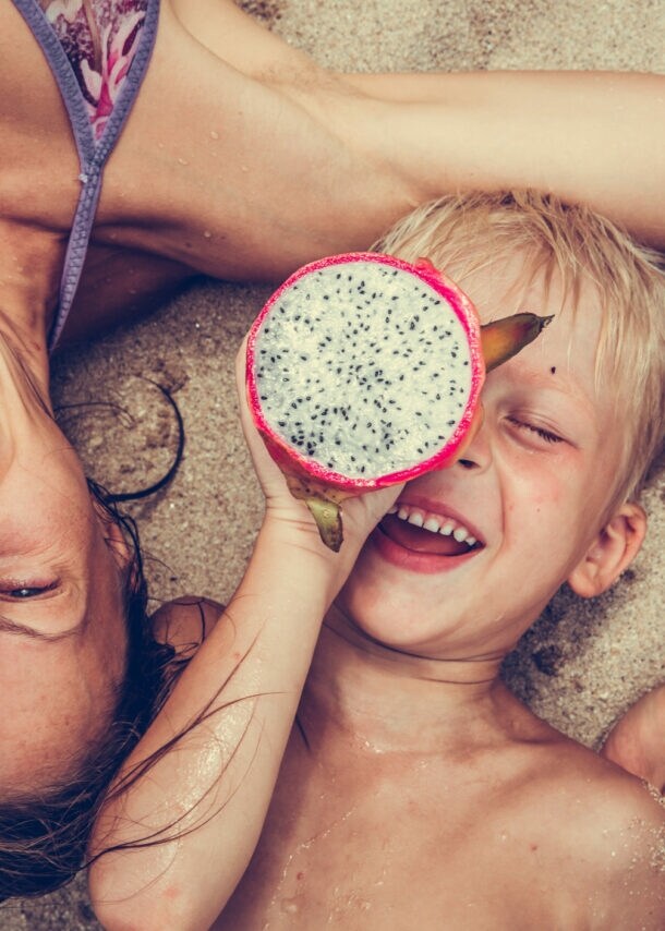 Eine Frau liegt mit einem Kind im Sand, beide halten eine tropische Frucht vor einem ihrer Augen.