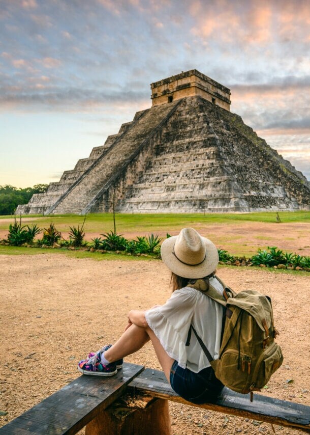 Person mit Hut und Rucksack sitzt auf einer Bank und blickt auf die Pyramide von Chichén Itzá bei Sonnenuntergang
