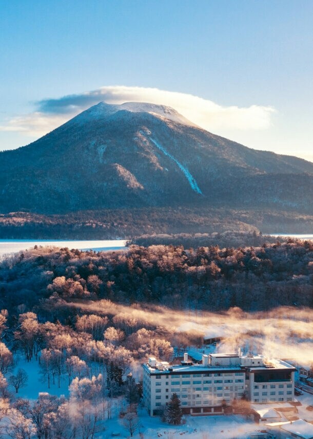 Verschneite Landschaft mit Wald und Gebäuden vor einem großen Berg unter klarem Himmel bei Sonnenaufgang