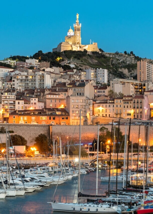 Abendliche Hafenansicht von Marseille mit Segelbooten im Wasser und der beleuchteten Kirche Notre-Dame de la Garde auf einem Hügel im Hintergrund