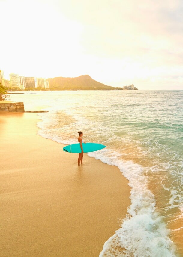 Eine Frau mit türkisfarbenem Surfboard unterm Arm steht am frühen Morgen am leeren Strand von Waikiki.