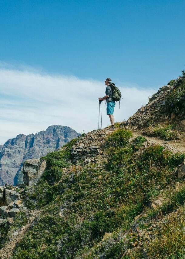Wanderer mit Rucksack und Wanderstöcken steht auf einem Bergpfad und blickt auf eine Berglandschaft unter blauem Himmel