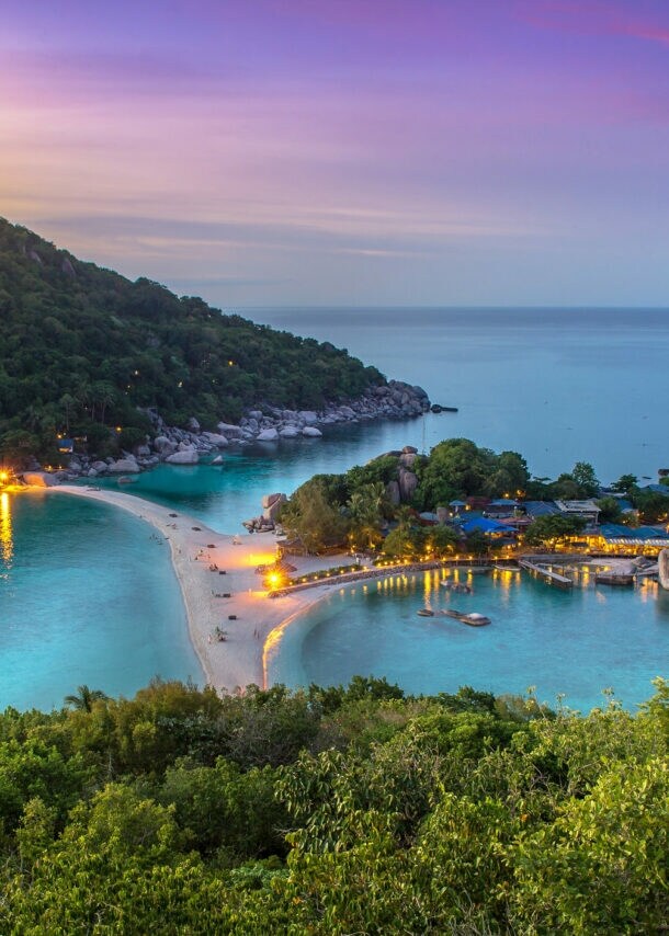 Blick auf eine tropische Insel mit bewaldetem Hügel, Sandbank und beleuchteten Gebäuden bei Sonnenuntergang