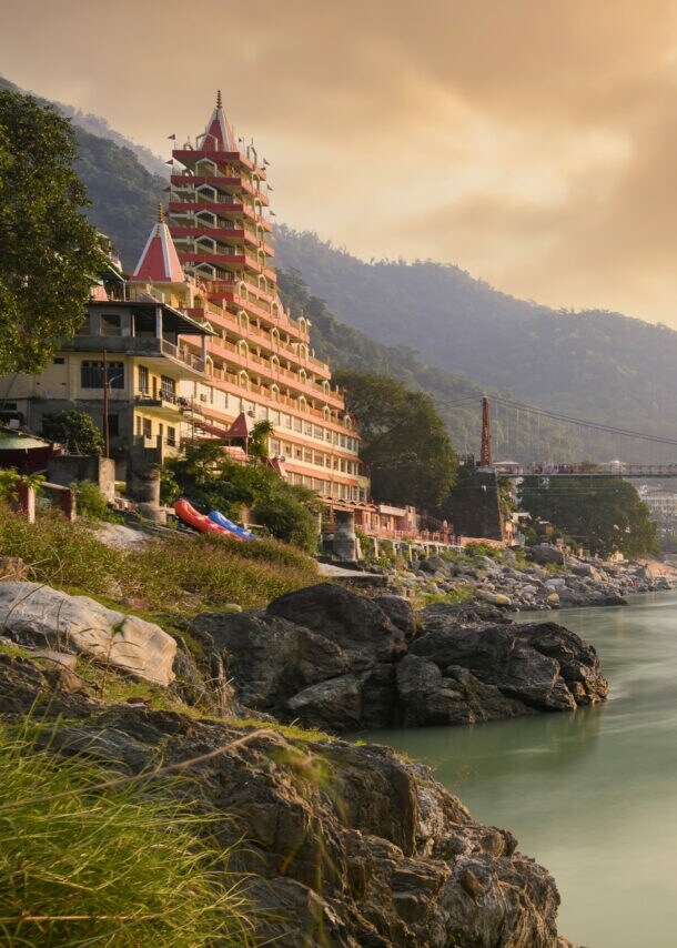 Fluss mit hängender Fußgängerbrücke und mehrstöckigem Tempelgebäude am Ufer bei Sonnenuntergang in bergiger Landschaft