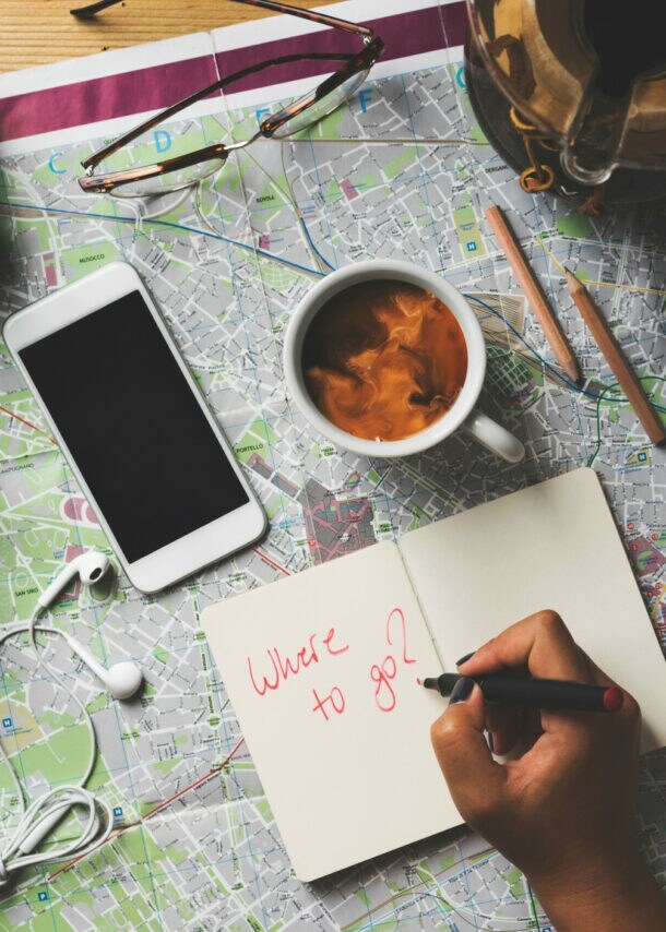 Hand schreibt 'Where to go?' in Notizbuch auf Landkarte mit Kamera, Smartphone, Kaffeetasse, Brille, Reisepass und Münzen