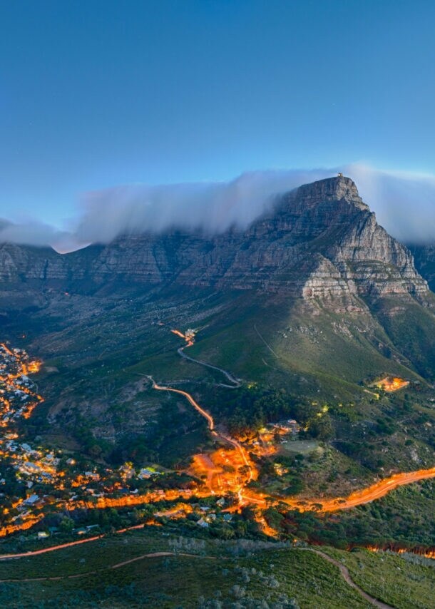 Tafelberg mit Wolkenkappe über Kapstadt bei Sonnenuntergang, beleuchtete Stadt und Küste im Hintergrund