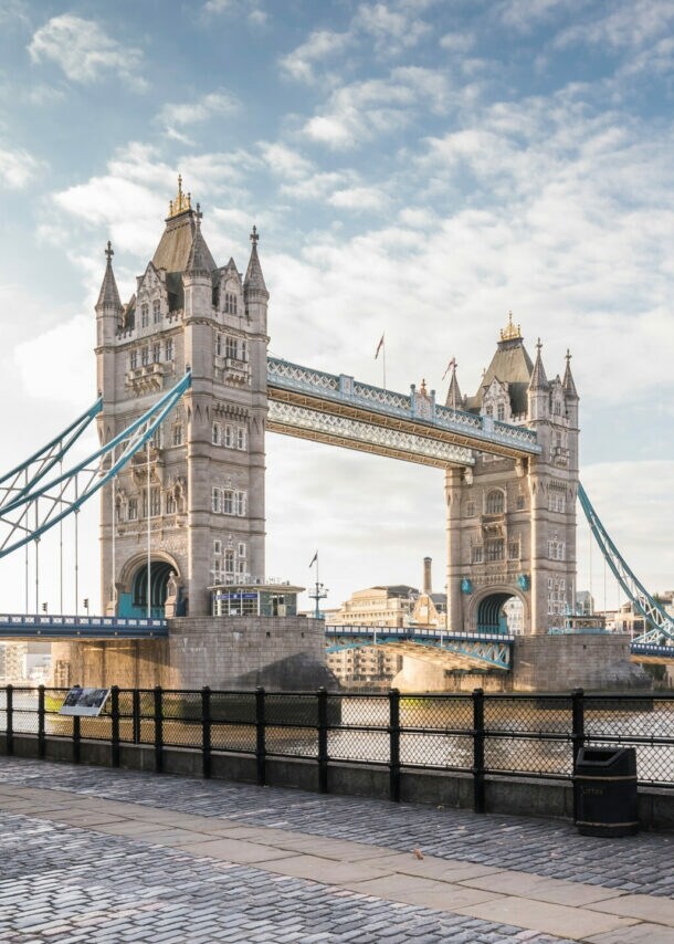 Tower Bridge über der Themse mit zwei markanten Türmen und blau-weißen Hängebrückenabschnitten, Blick vom Ufer mit Bäumen und Laterne.