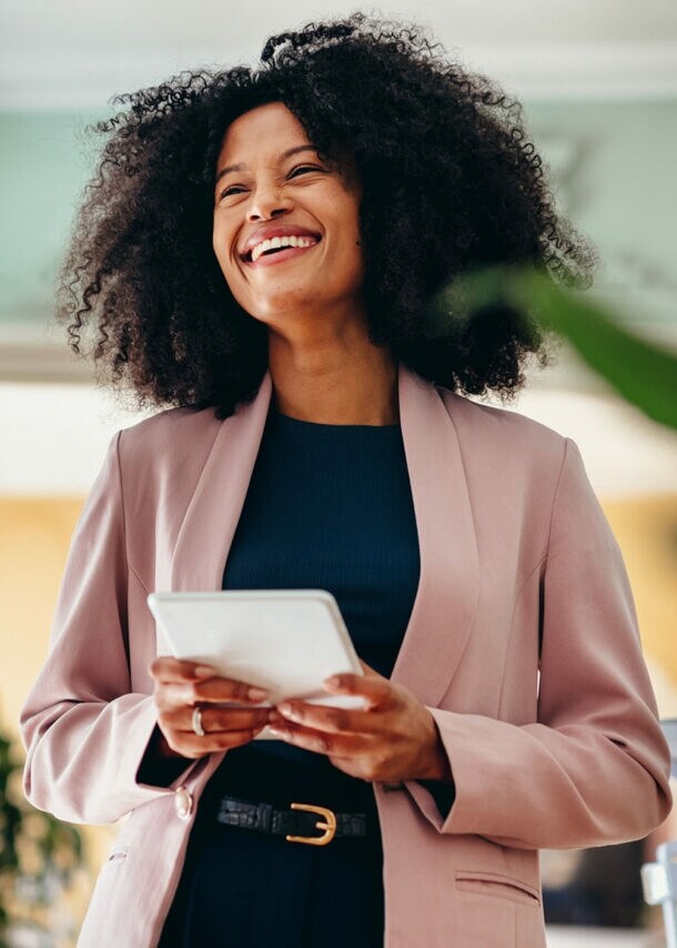 Eine lachende Frau in einem rosafarbenen Blazer steht in einem modernen Büro und hält ein weißes Tablet in den Händen, daneben ein Freisteller einer silbernen American Express Business Kreditkarte über einer einfachen American Express Platinum Card.