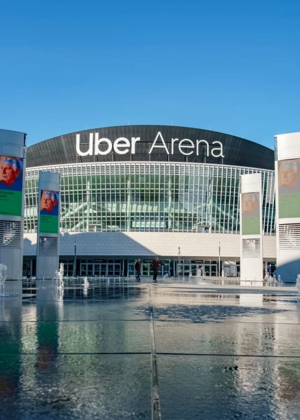 Rundes Gebäude mit Glasfassade und der Aufschrift Uber Arena hinter einem bebauten Platz mit Werbetafeln um eine Wasserfläche.