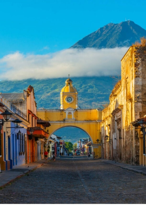 Blick auf die gelbe Santa-Catalina-Arkade in Antigua, Guatemala, mit Kopfsteinpflasterstraße und Vulkan Agua im Hintergrund