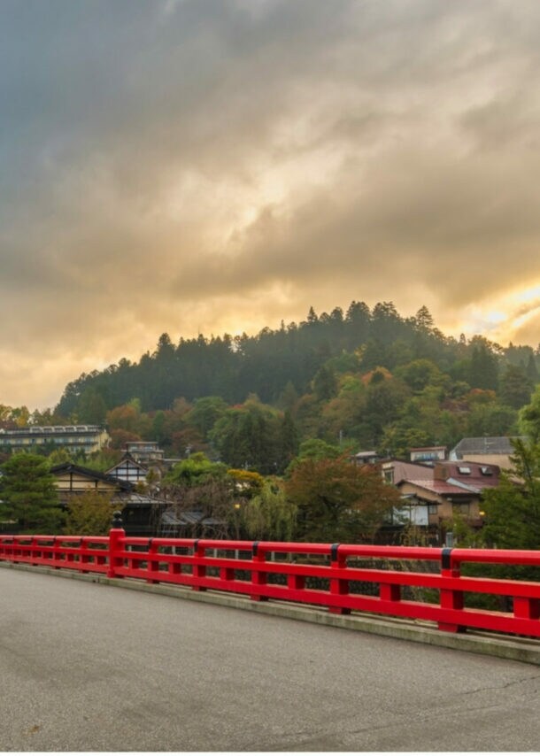 Rote Brücke über Fluss mit Häusern und bewaldetem Hügel im Hintergrund bei bewölktem Himmel