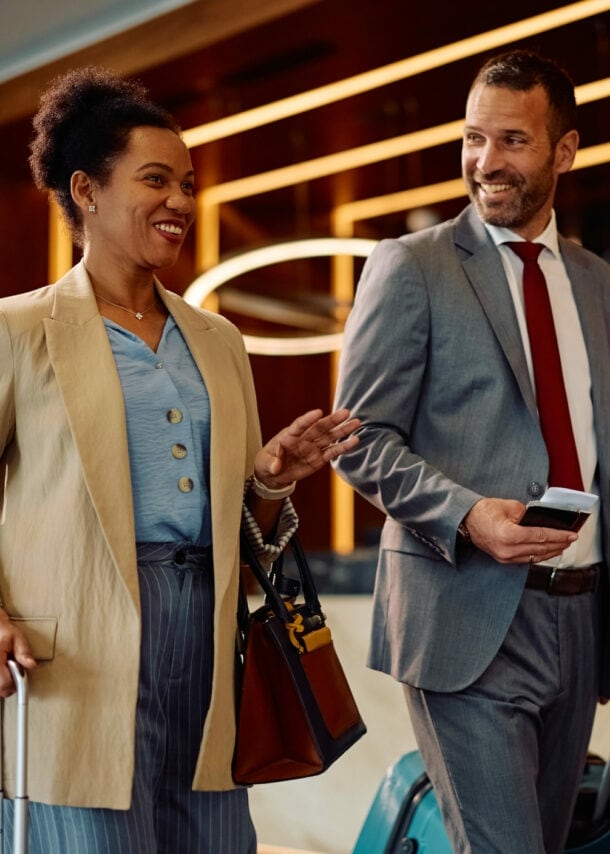 Eine Frau und ein Mann in Business-Kleidung mit Trolleys gehen gut gelaunt durch eine Hotellobby.