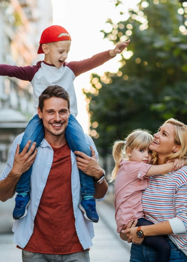 Ein Mann auf einem Weg zwischen Haus und Straße trägt einen Jungen auf den Schultern, eine Frau daneben trägt ein Mädchen.