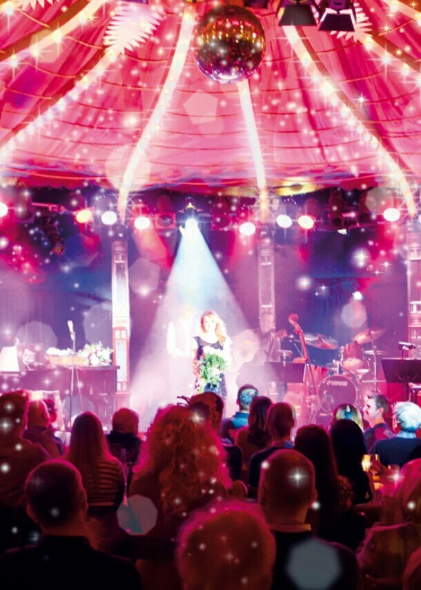 Bühne im Bar jeder Vernunft Theater Berlin mit Künstlerin im Scheinwerferlicht vor Publikum unter Zelt mit Lichterketten