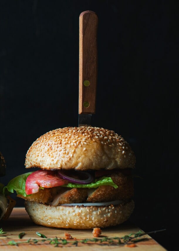 Drei verschiedene Burger mit Messern im Brötchen auf einem Holzbrett, darunter Salat, Tomate und Käse sichtbar