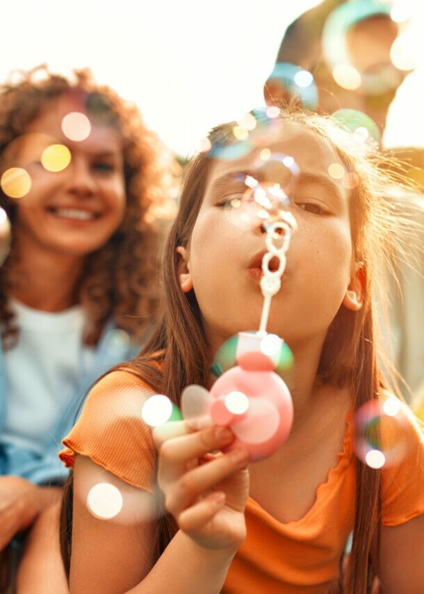 Eltern mit Kindern sitzen draußen auf Gras und spielen mit Seifenblasen bei Sonnenlicht.
