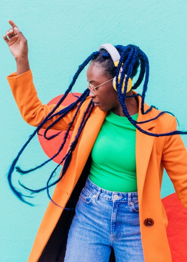 Eine junge Frau mit blauen Rastazöpfen in einem grünen Shirt und orangefarbenen Mantel tanzt mit gelben Kopfhörern vor einer türkisfarbenen Fassade.