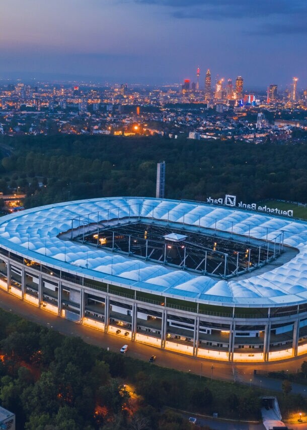 Luftaufnahme eines Fußballstadions, umgeben von Waldgebiet, vor der erleuchteten Skyline Frankfurts bei Nacht.