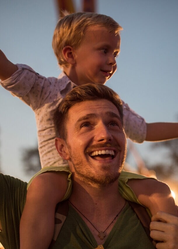 Mann im Dunkeln mit kleinem Kind auf den Schultern, im Hintergrund sind Bäume und Beleuchtung zu sehen.