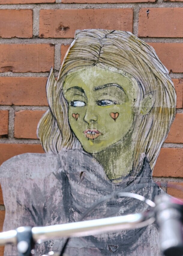 Frau mit braunen Haaren und rotem Blumenmuster-Oberteil fotografiert eine Wandzeichnung einer Person mit langen Haaren vor einer Backsteinmauer.