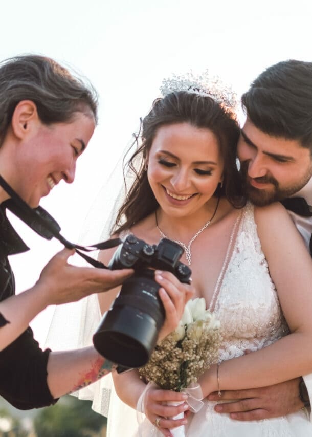 Braut in weißem Spitzenkleid mit Blumenstrauß, Bräutigam in weißem Anzug umarmt sie von hinten, Fotografin zeigt Kamera.