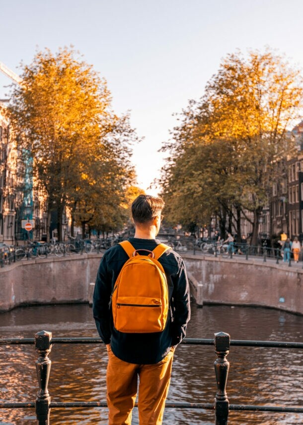 Rückansicht einer Person mit Rucksack, die an einem sonnigen Tag an einem Kanal in Amsterdam steht, auf der anderen Seite Häuser.