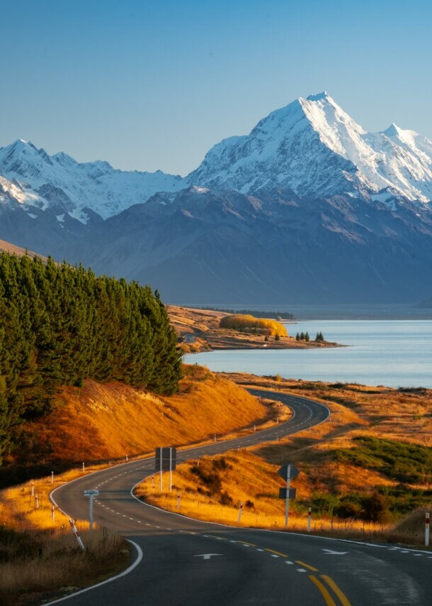 Malerischer Blick auf schneebedeckte Berge vor klarem blauem Himmel, im Vordergrund verläuft eine verlassene Straße zwischen grünen Nadelwäldern und einem ruhigen See.