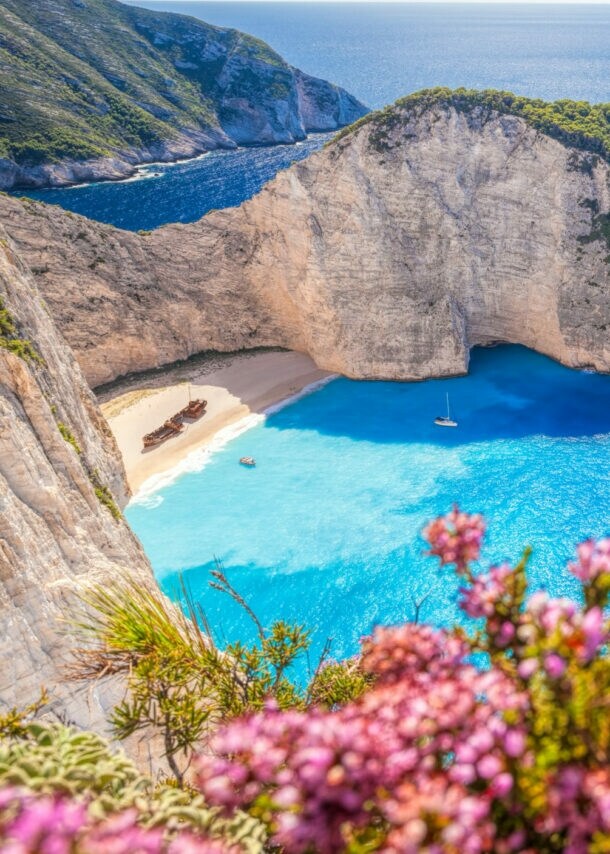 Bucht mit weißem Sandstrand und gestrandeten Schiffsrumpf, umgeben von steilen Klippen und blauem Meer, violette Blumen im Vordergrund
