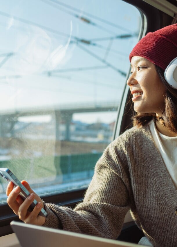 Eine Person mit Kopfhörer und Smartphone sitz in einem Zug. Sie trägt eine rote Mütze und einen grauen Pullover. Im Hintergrund sind Züge und Schienen zu sehen.
